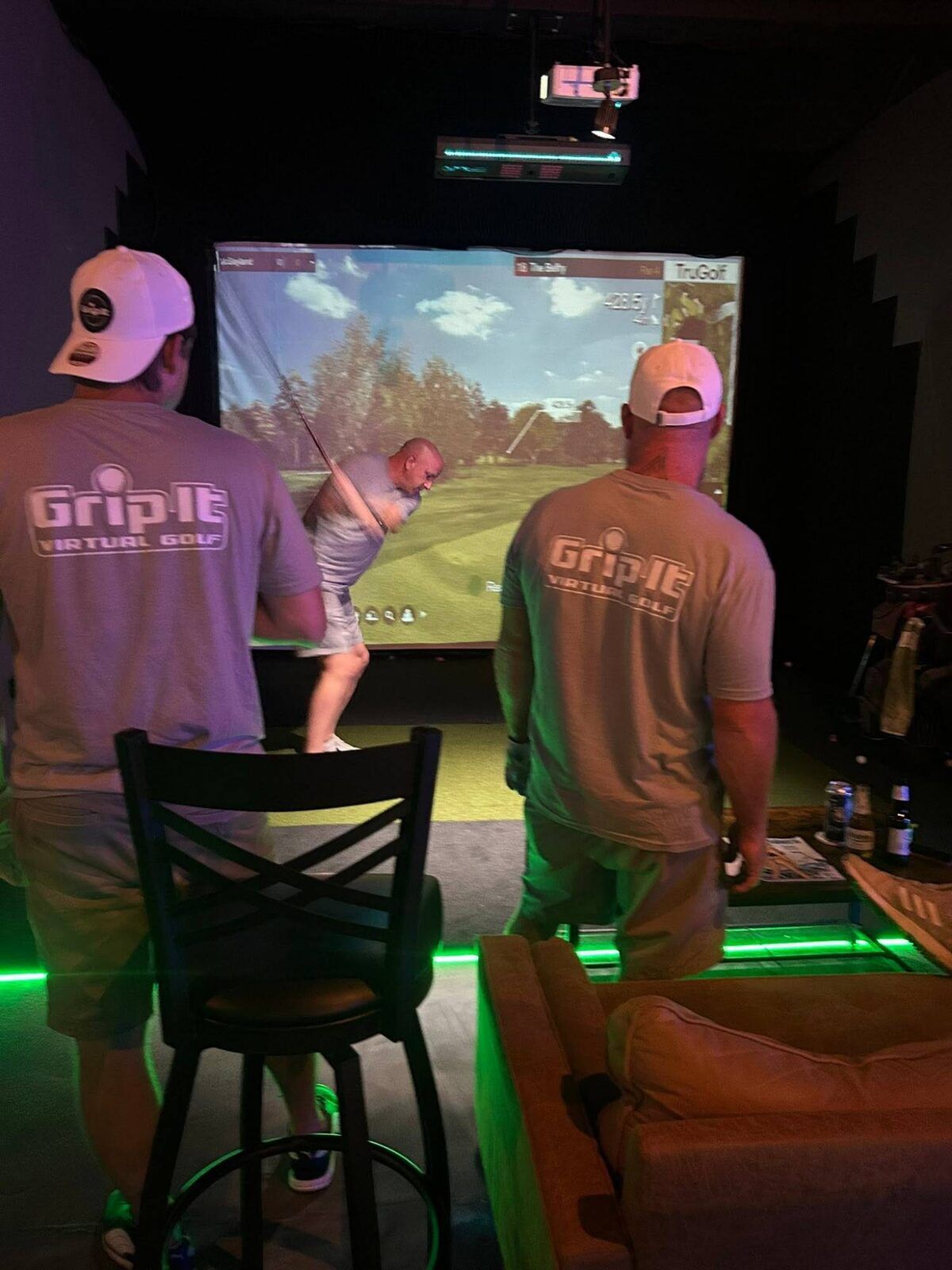 A golfer mid-swing on the simulator at Grip It Virtual Golf while two friends in Grip It shirts watch